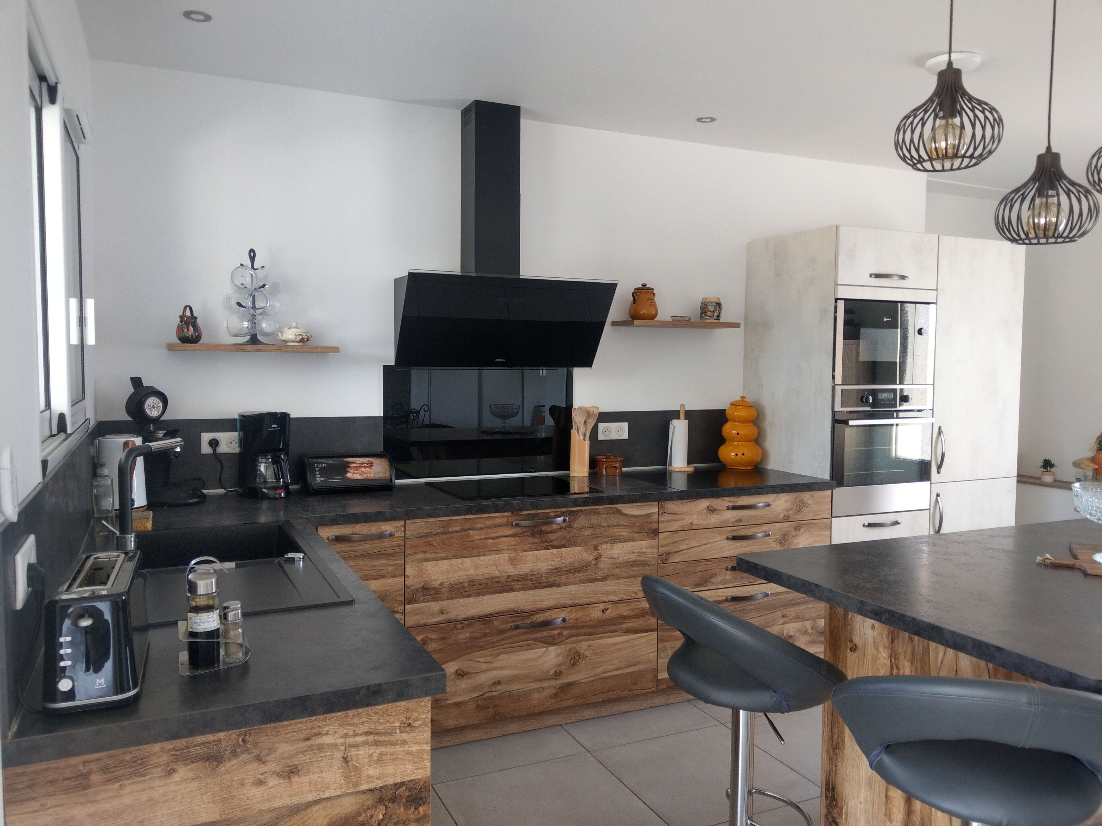 Une cuisine moderne dotée d’une hotte noire, de meubles en bois, d’un four encastré et d’un coin bar avec des tabourets.