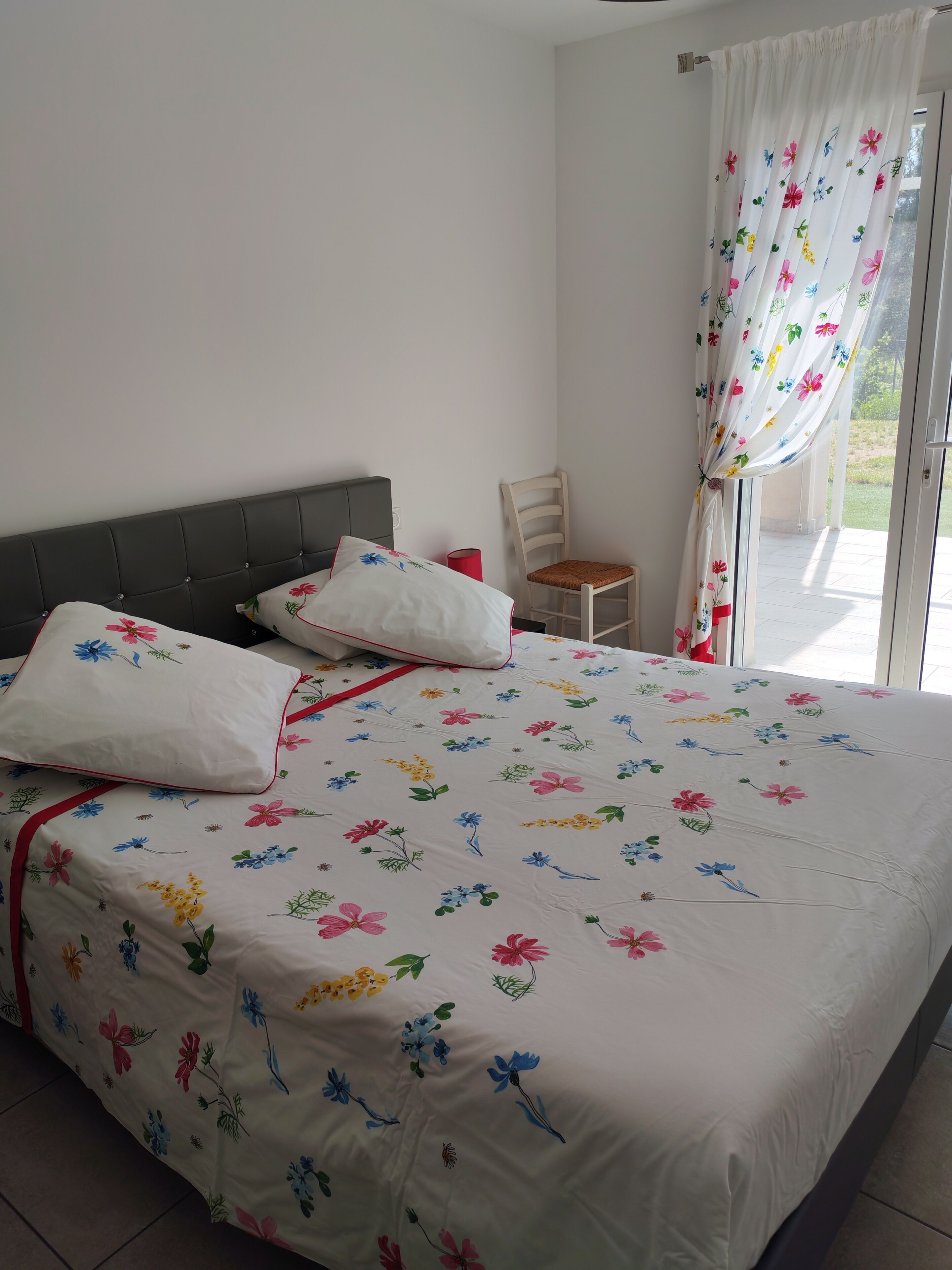 Un lit recouvert d’une couvre-lit à motifs floraux, une chaise en bois et une fenêtre avec des rideaux.