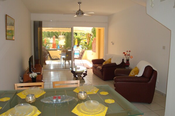 Uma sala de jantar com mesa de vidro posta para quatro pessoas, um sofá de couro marrom e vista para um pátio com móveis de exterior.