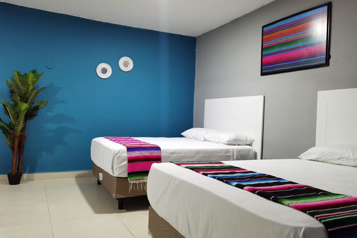 Quarto com duas camas, parede azul decorativa, planta e obra de arte colorida.