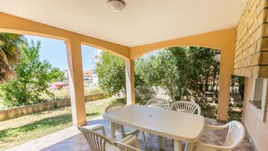 Un porxo cobert amb una taula i cadires, un paviment de rajola i vista a la vegetació i edificis al fons.