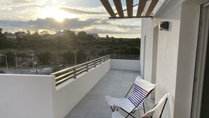 A balcony with two white chairs, a railing, and a view of a road and buildings in the distance.