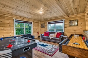 A cozy room with wooden walls, a pool table, a sofa with red cushions, and a large window.
