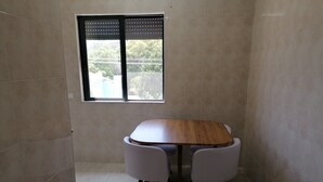 Een kleine, betegelde kamer met een houten tafel en vier stoelen. Er is een raam met een metalen kader en uitzicht op groen buiten.