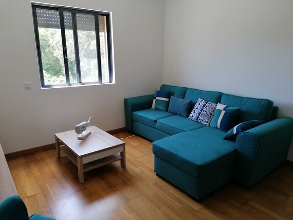 Ein Wohnzimmer mit einem blaugrünen Sofa, einem kleinen Holztisch und einem Fenster mit Jalousien.