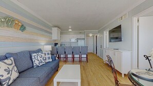 Un ambiente compatto con divano, angolo da pranzo con sedie, zona cucina con pensili ed elettrodomestici, e televisore montato a parete.