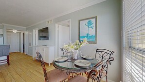 Un'area pranzo con tavolo in vetro, sedie bianche e un vaso con fiori. Si nota un quadro appeso al muro e una finestra con tende.