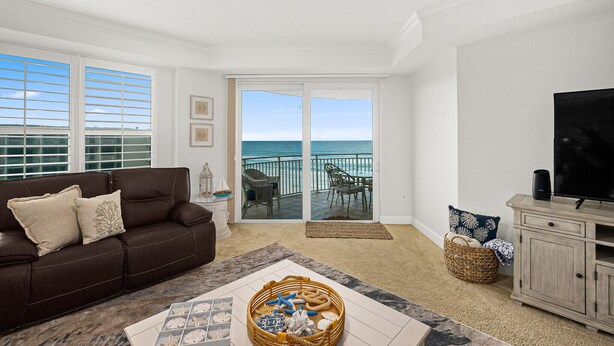 A living room with a brown leather sofa, a coffee table with decorative items, a television on a wooden cabinet, and a view of the ocean through a sliding glass door.