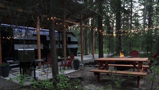 An RV parked under a wooden pavilion with string lights, surrounded by trees and a picnic table with a fire pit.