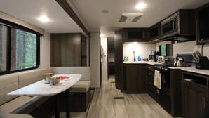 A compact kitchen with dark cabinets, a microwave, and a stove. There is a window with a view of trees, and a bench with a table in the foreground.