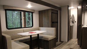 A compact RV interior with a cushioned seating area, a small table, and a window offering a forest view.
