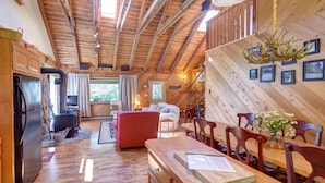 Interior rústico de una cabaña con techo de madera, un comedor con sillas de madera, un sofá rojo y una chimenea.