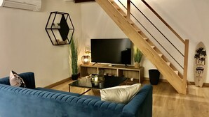 A living room with a blue sofa, a coffee table, a TV, and a staircase. - Les Cocons du Bassin - Cocon Duplex&design - Clim/wifi/linge Compris (Gujan-Mestras)