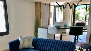 A modern living room with a blue sofa, a glass coffee table, and a view of a dining area through a glass door. - Les Cocons du Bassin - Cocon Duplex&design - Clim/wifi/linge Compris (Gujan-Mestras)