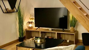 A modern living room with a flat-screen TV, wooden staircase, and a surfboard decoration. - Les Cocons du Bassin - Cocon Duplex&design - Clim/wifi/linge Compris (Gujan-Mestras)