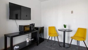 Une petite pièce avec un téléviseur à écran plat fixé au mur, un meuble noir pour la télévision avec un micro-ondes, un petit réfrigérateur et deux chaises jaunes disposées autour d’une table ronde sur laquelle se trouve un pot de fleurs.