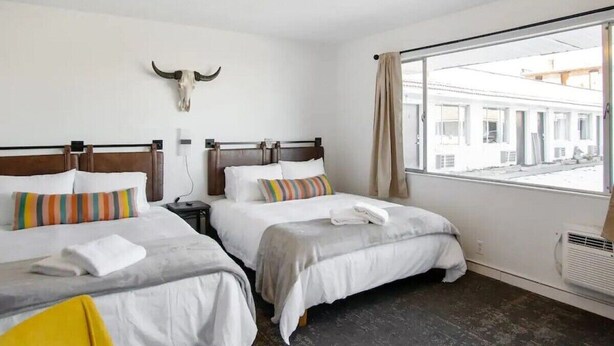 Une chambre d’hôtel avec deux lits, une grande fenêtre donnant sur des immeubles, un décor mural représentant un crâne de vache et un lustre moderne.
