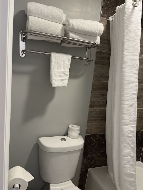 Et badeværelse med toilet, et håndklædestativ med foldede håndklæder, toiletpapir og en bruseforhæng.