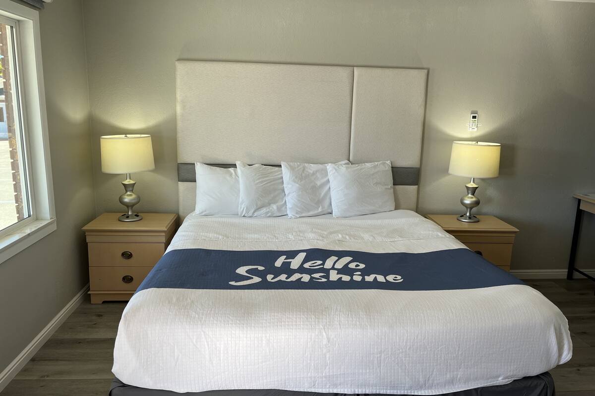 A neatly made bed with a 'Hello Sunshine' blanket, two bedside lamps, and a window on the left side.