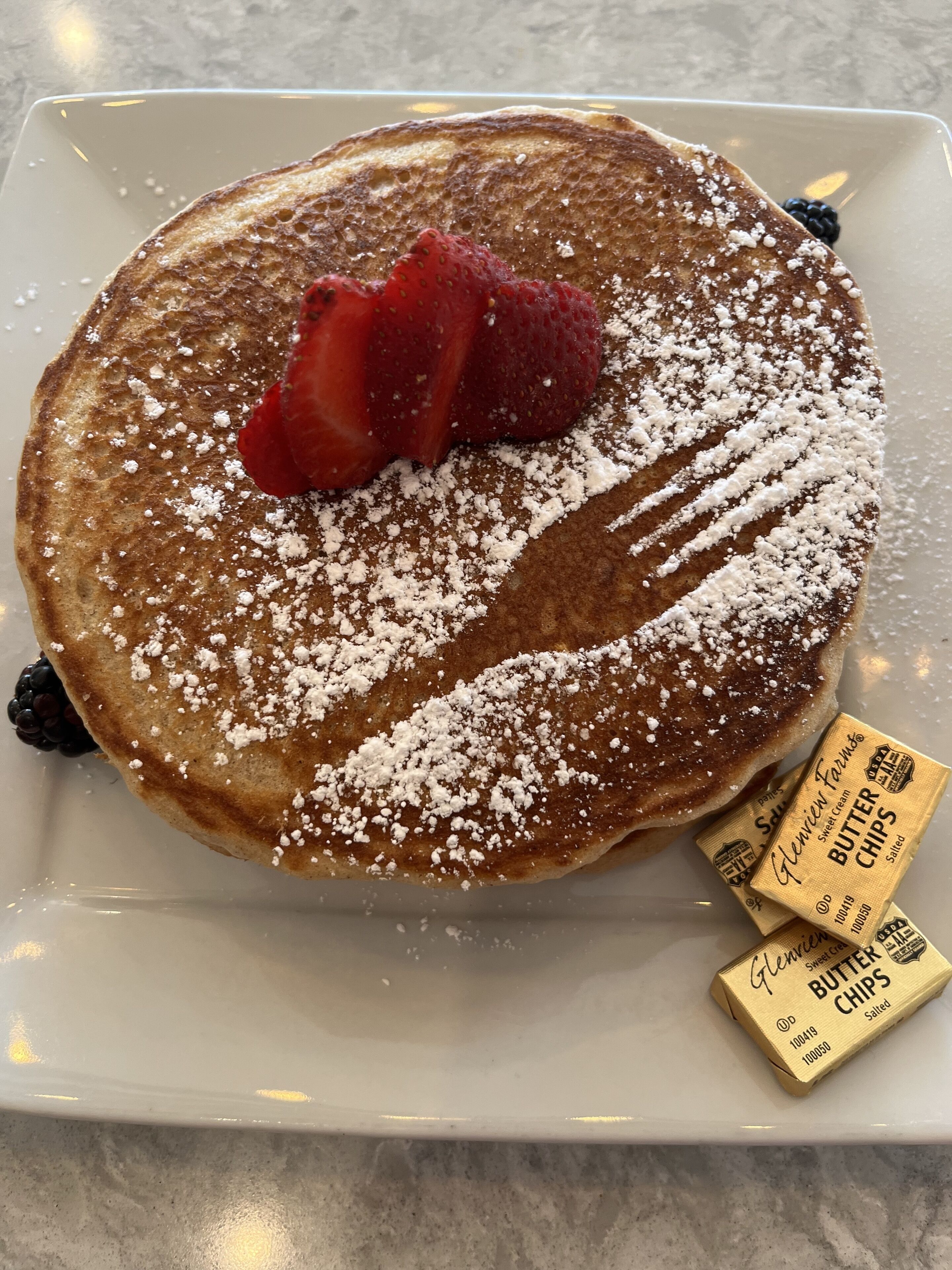 A plate of pancakes with powdered sugar and fresh berries, accompanied by a box of salted butter chips.