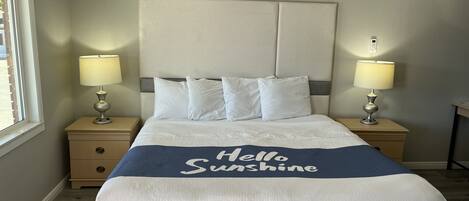 Ein ordentlich bezogenes Bett mit einer „Hello Sunshine“-Bettdecke, zwei Nachttischlampen und einem Fenster auf der linken Seite.
