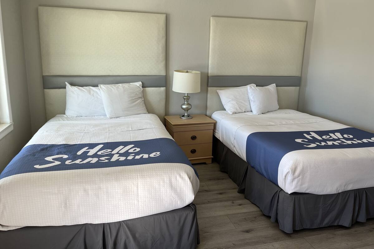 A hotel room with two beds, each with a 'Hello Sunshine' bedspread, a bedside lamp, and a nightstand with a drawer.
