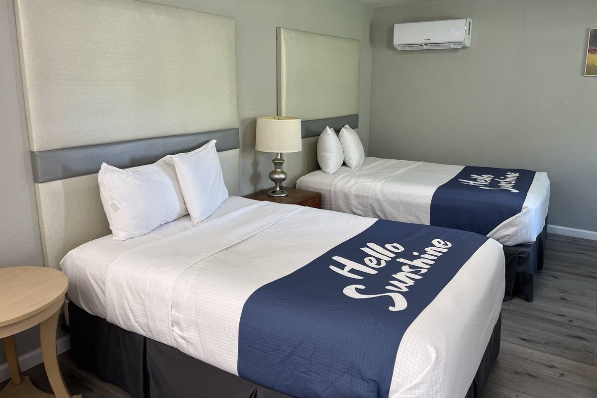 A hotel room with two beds, each with a 'Hello Sunshine' bedspread, a bedside table with a lamp, and an air conditioning unit on the wall.