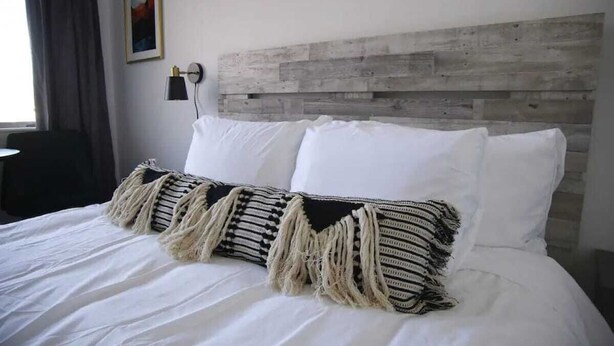 A bed with white linens and a striped throw pillow, a wooden headboard, and a wall-mounted lamp.