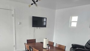 A modern dining area with a wooden table, black chairs, a TV mounted on the wall, and a ceiling light fixture.