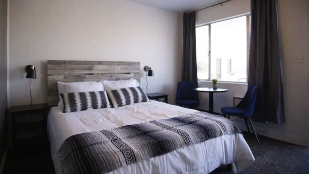 A hotel room with a large bed, a headboard, two bedside tables, a small round table, two chairs, and a large window with curtains.