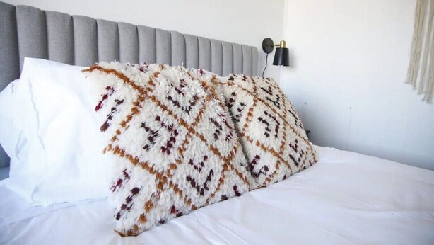 Een bed met een gebogen deken, een grijs gestoffeerd hoofdbord en een wandlamp.