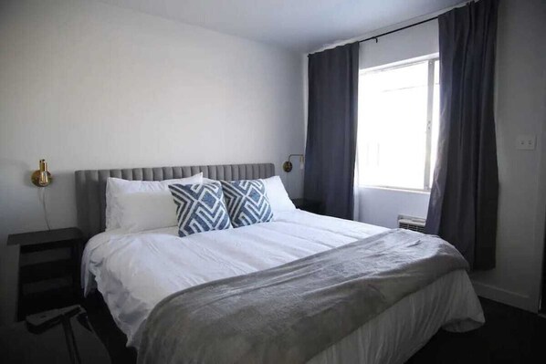 A hotel room with a large bed, a bedside table, a lamp, and a window with curtains. - Fillmore Lodge: One King Room with Lowest Rates! (Fillmore)