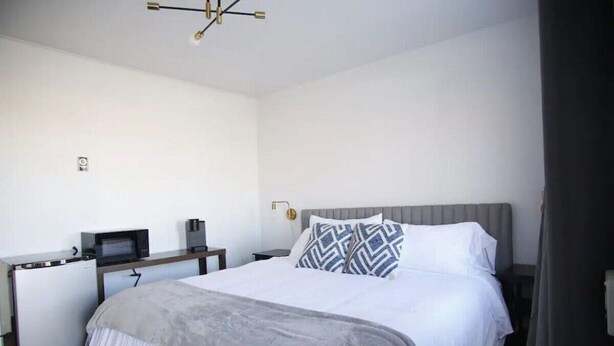 A hotel room with a bed, a desk with a microwave, a small refrigerator, and a bedside table with a lamp. - Fillmore Lodge: One King Room with Lowest Rates! (Fillmore)