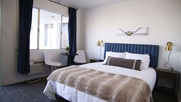 Quarto de hotel com uma cama grande, uma escrivaninha com cadeira, uma janela com cortinas e um aplique de parede.