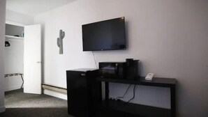 Quarto com televisão de tela plana na parede, escrivaninha preta com um computador e um cacto decorativo.