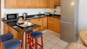 A kitchen with wooden cabinets, a stainless steel refrigerator, a microwave, a kettle, and a coffee maker. There are two bar stools and a wicker chair. - La Montagne (Ballito)