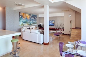 A modern living room with a white sofa, a large painting, a flat-screen TV, and a dining area with purple chairs and a glass table.