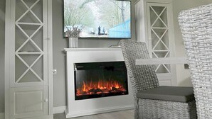 A living room with a fireplace, a television mounted above it, and a wicker chair. - Rugiamare, Exclusives, neu Renoviertes, Strandnahes Reihen- Ferienhaus (Dranske)