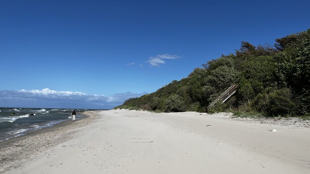 A beach with a person walking along the shore, a staircase leading up to a wooded area, and the ocean in the background. - Rugiamare, Exclusives, neu Renoviertes, Strandnahes Reihen- Ferienhaus (Dranske)