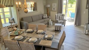 A dining area with a table set for six, surrounded by wicker chairs. A sofa and striped armchairs are visible in the background. - Rugiamare, Exclusives, neu Renoviertes, Strandnahes Reihen- Ferienhaus (Dranske)