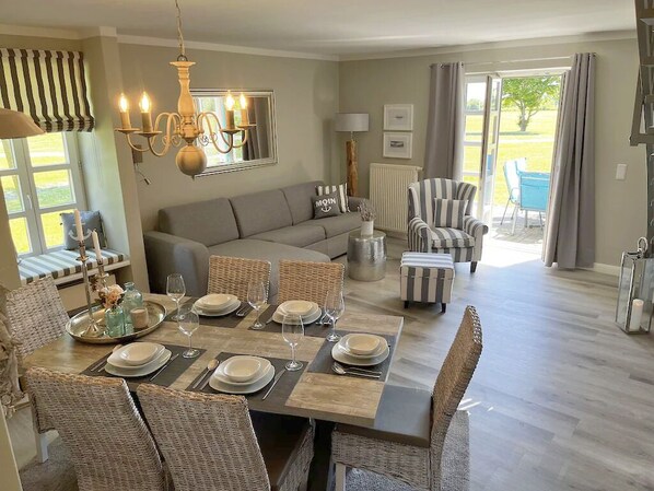 A dining area with a table set for six, surrounded by wicker chairs. A sofa and striped armchairs are visible in the background.