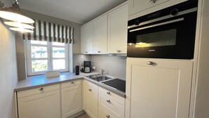 A modern kitchen with white cabinets, a black oven, and a window with striped curtains. - Rugiamare, Exclusives, neu Renoviertes, Strandnahes Reihen- Ferienhaus (Dranske)