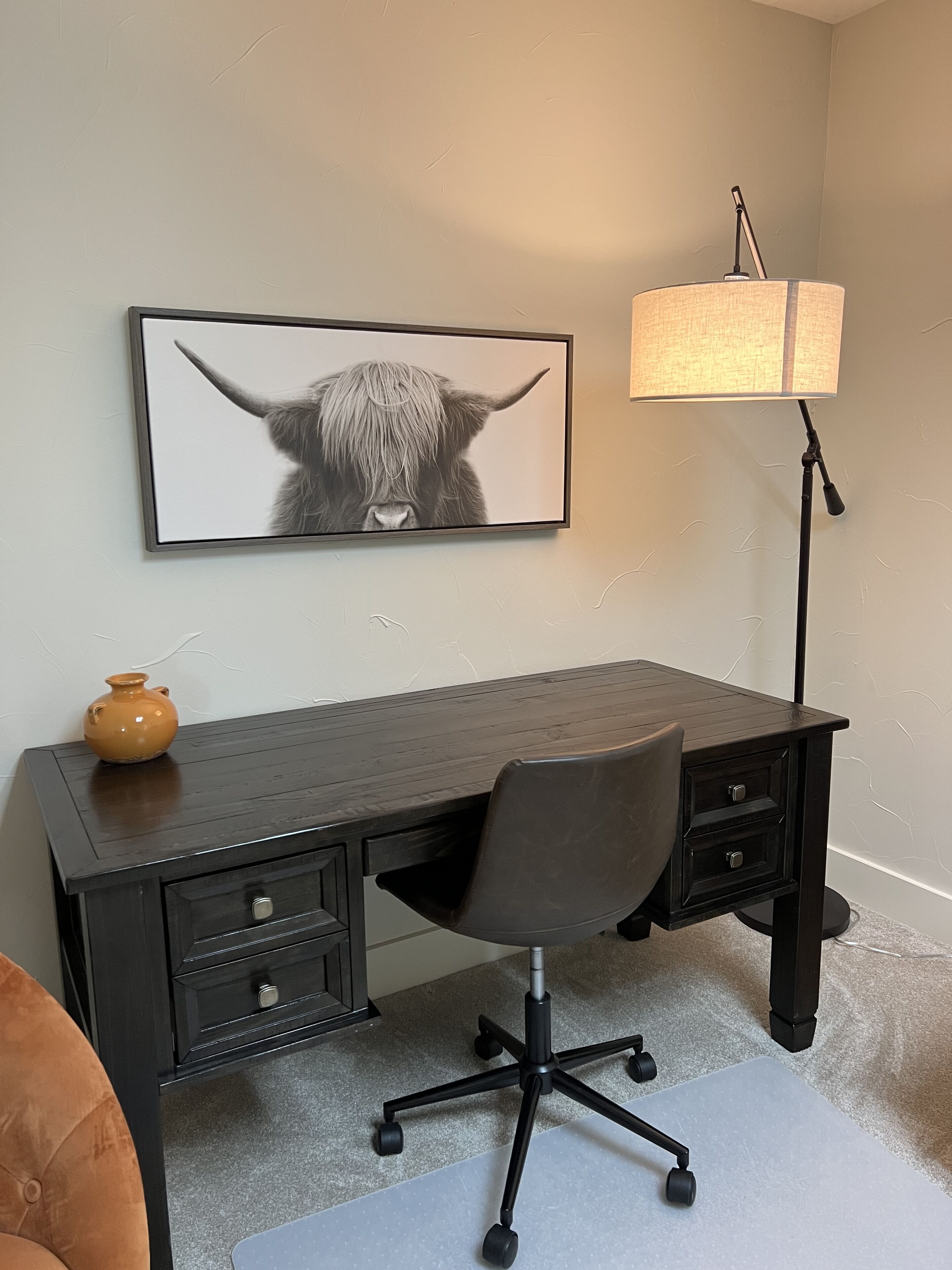 Ein dunkler Holztisch mit einem schwarzen Bürostuhl, ein gerahmtes Tierbild an der Wand, eine Stehlampe und ein kleiner Keramiktopf auf dem Tisch.
