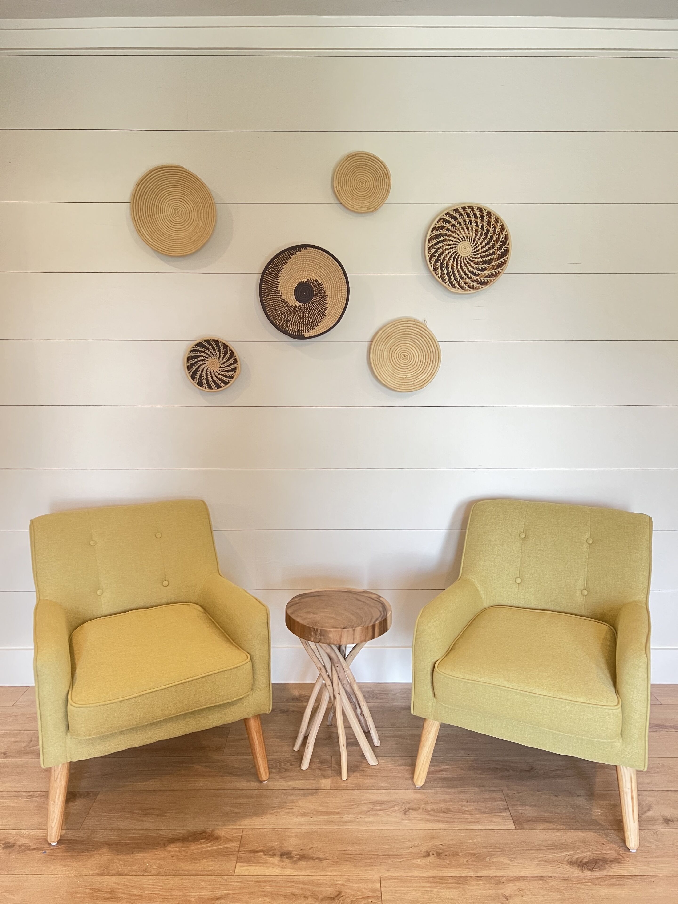 A room with two yellow armchairs, a small wooden side table, and several woven wall decorations.