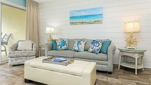 Una sala de estar con un sofá gris, un otomano color crema y una mesita de mimbre. En la pared, hay un cuadro de una playa.