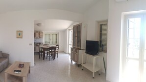 A compact living space with a sofa, a small table, a TV on a stand, and a kitchen in the background. - The Riace Palms 3 (Riace Marina)