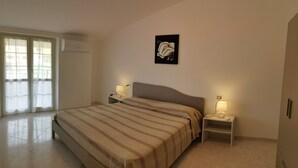 A bedroom with a bed, two bedside tables with lamps, a wall-mounted air conditioner, and a window with curtains. - The Riace Palms 3 (Riace Marina)