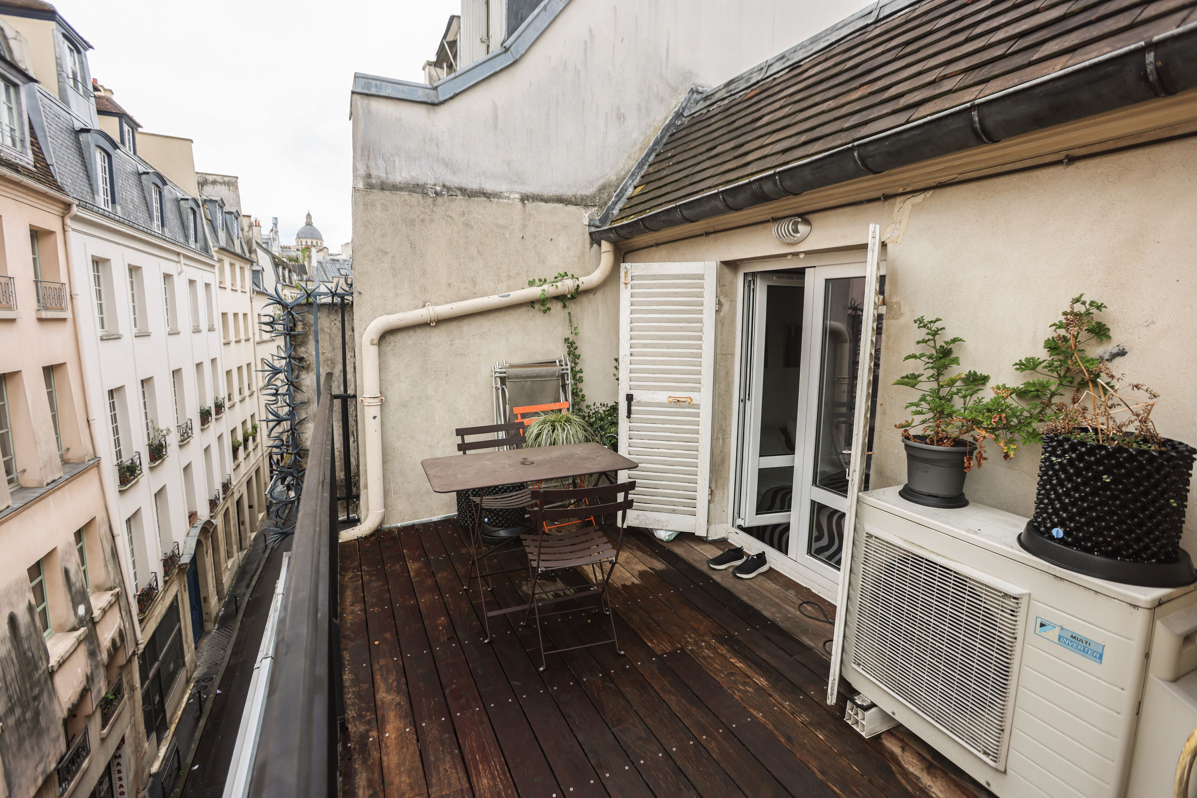 A narrow balcony with a wooden table and chairs, a potted plant, and a window with shutters.