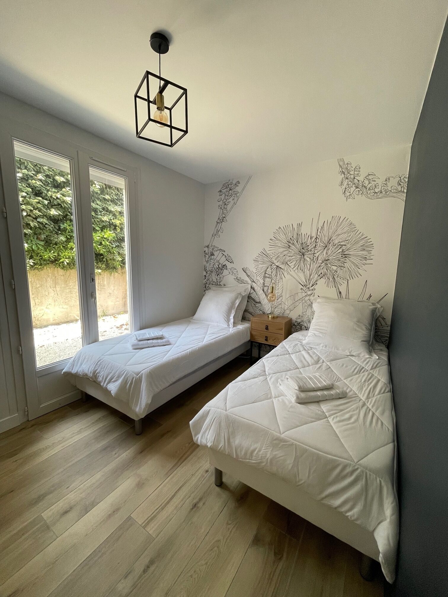 A bedroom with two single beds, a wooden floor, a ceiling light fixture, and a window with a view of greenery.