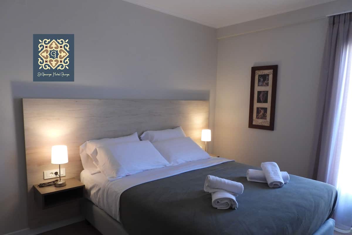 A neatly made bed with white linens and two bedside lamps, a framed picture on the wall, and a logo for St. George Hotel Parga in the top left corner.